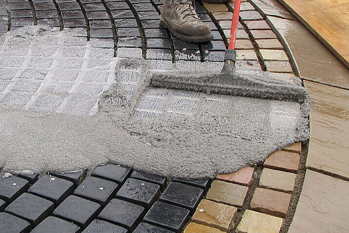 Worker applying joint grout to curved stone paver patio using a push broom